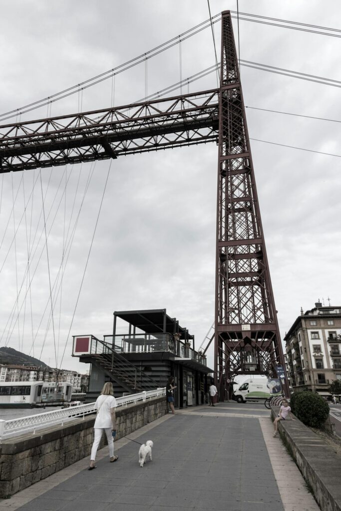 A woman walks her dog near a historic red iron bridge on a cloudy day.